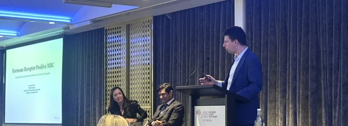 Dr Joshua Hurwitz presenting on hormone receptor-positive metastatic breast cancer at the Breast Cancer Trials conference, 2026, with panel members seated beside the stage